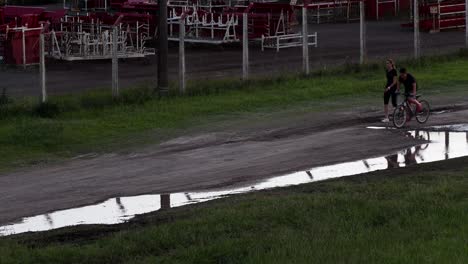 A-woman-walks-with-a-child-on-a-bycicle-over-a-muddy-road,-their-reflection-seen-on-a-puddle