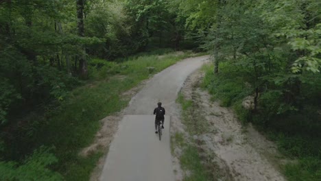 Drone-Siguiendo-A-Un-Hombre-Haciendo-Ejercicio-Andando-En-Bicicleta-Por-Un-Tranquilo-Sendero-Para-Bicicletas-En-El-Bosque-Vestido-Con-Un-Traje-Negro