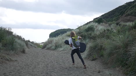 Un-Joven-Rubio-Corriendo-Alegremente-Hacia-El-Océano,-Con-Una-Mochila-Y-Una-Funda-De-Guitarra,-En-Un-Sendero-Arenoso-Rodeado-De-Hierba-Alta-En-La-Playa-De-Bethells,-En-Un-Día-Nublado-Y-Ventoso