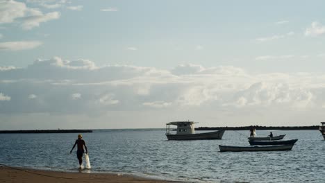 Fisherman-holding-fishing-net-walking-out-of-the-sea-in-slow-motion-on-a-sunny-day-with-boats-on-the-horizon