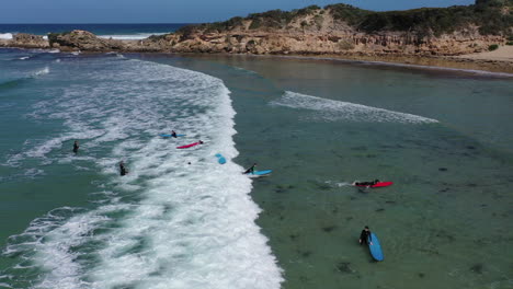 Aéreo:-Un-Estudiante-Sobre-Una-Tabla-De-Surf-Azul-Se-Lanza-A-Una-Pequeña-Rompiente-En-La-Costa