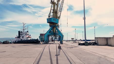 Aerial-view-of-a-woman-walking-towards-a-large-blue-shipping-crane-on-a-shipping-dock-,-tracking-wide-shot