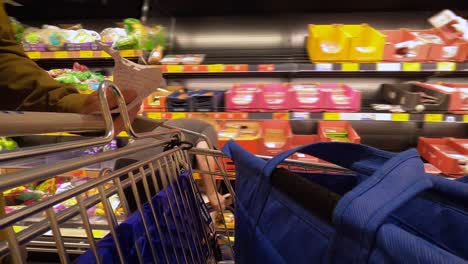 Man-pushing-a-shopping-cart-along-a-busy-aisle-of-random-people-past-fresh-fruit-and-vegetables-then-refrigerated-meats-while-holding-a-food-list