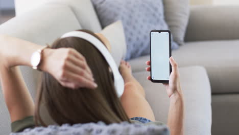 A-Caucasian-woman-is-holding-a-smartphone-with-a-blank-screen-on-a-couch-at-home,-copy-space