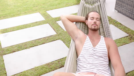 A-young-Caucasian-male-relaxes-in-a-hammock-in-the-backyard-at-home,-eyes-closed,-copy-space