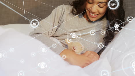 Animation-of-network-of-connections-with-icons-over-biracial-woman-lying-on-bed-with-pet-cat