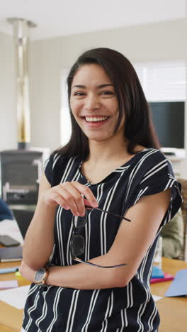 Retrato-En-Video-Vertical-De-Una-Mujer-Birracial-Sonriente,-Colegas-Diversos-En-El-Fondo,-Cámara-Lenta