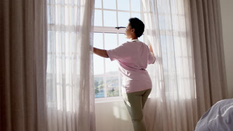 African-American-woman-opens-curtains-to-a-sunlit-room-at-home