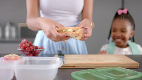 Una-Joven-Madre-Birracial-Prepara-Un-Sándwich-En-Casa,-Con-Una-Hija-Afroamericana-Sonriendo-Cerca