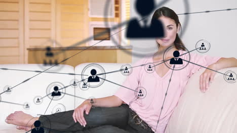 Animation-of-network-of-connections-with-icons-over-caucasian-woman-sitting-on-sofa