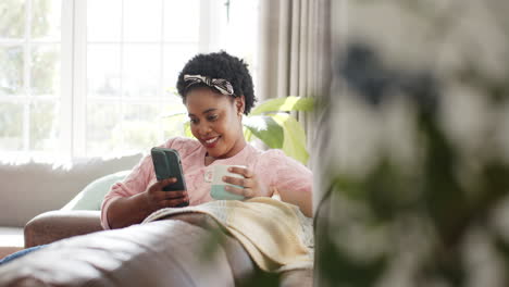 African-American-woman-smiles-at-her-phone,-holding-a-mug-at-home