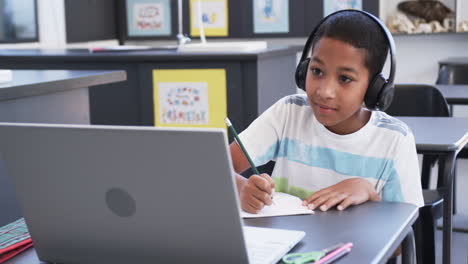 Biracial-boy-with-headphones-uses-a-laptop-and-writes-notes-in-a-classroom-at-school