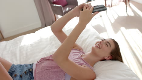 Kaukasisches-Teenager-Mädchen-Mit-Braunen-Haaren-Nutzt-Ein-Smartphone,-Während-Sie-Zu-Hause-Auf-Einem-Bett-Liegt