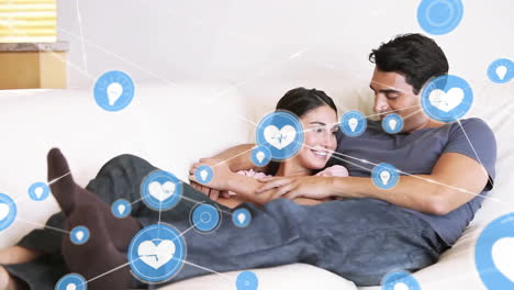 Animation-of-network-of-connections-with-icons-over-caucasian-couple-embracing-on-sofa