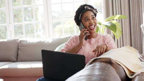 Una-Mujer-Afroamericana-Habla-Por-Teléfono-En-Casa,-Una-Computadora-Portátil-Abierta-Ante-Ella