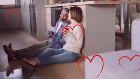 Animation-of-hearts-over-diverse-couple-drinking-coffe-and-sitting-on-floor-in-kitchen-at-home