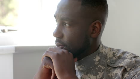 African-American-soldier-in-military-uniform-looks-pensive,-resting-chin-on-hand-at-home