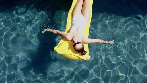 Teenage-Caucasian-girl-relaxes-on-a-yellow-float-in-a-pool,-wearing-sunglasses