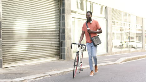 African-American-man-walks-with-his-bike,-holding-a-coffee-cup-with-copy-space-in-the-city