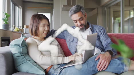 Animation-of-house-shape-over-diverse-couple-sitting-on-sofa-using-laptop-at-home