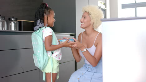 African-American-child-with-a-backpack-hands-a-gift-to-a-young-biracial-mother-in-a-kitchen