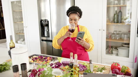 Una-Mujer-Afroamericana-Bebe-Vino-Y-Revisa-Su-Teléfono-En-Casa-Mientras-Cocina