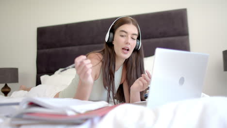 Teenage-Caucasian-girl-studies-with-a-laptop-in-bed-at-home,-wearing-headphones