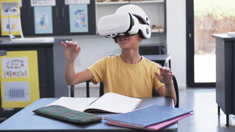 Biracial-boy-wearing-a-VR-headset-is-seated-at-a-desk-with-notebooks-in-a-classroom-at-school