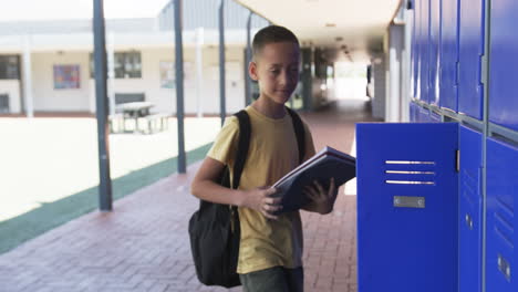 Biracial-boy-with-a-backpack-stands-in-a-school-corridor,-holding-textbooks