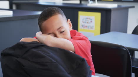 Biracial-child-with-freckles-in-red-shirt-rests-head-on-arms-in-school-classroom