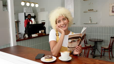 A-young-biracial-woman-with-curly-blonde-hair-smiles-while-reading-a-book-at-a-cafe