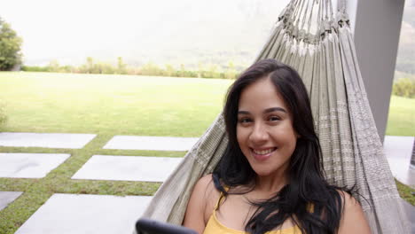 A-young-biracial-woman-lounges-in-a-hammock-in-the-backyard-at-home,-smartphone-in-hand,-copy-space
