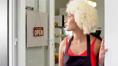 Young-biracial-woman-with-curly-blonde-hair-stands-by-an-open-cafe-door