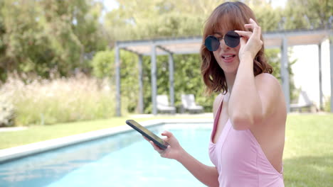 Young-biracial-woman-in-a-pink-swimsuit-smiles-while-using-her-phone-by-the-pool-with-copy-space