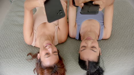 Two-young-biracial-female-friends-lie-back-to-back,-smiling-while-holding-smartphones