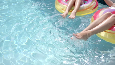 Young-women-splash-water-with-their-feet-while-sitting-on-colorful-pool-floats,-with-copy-space