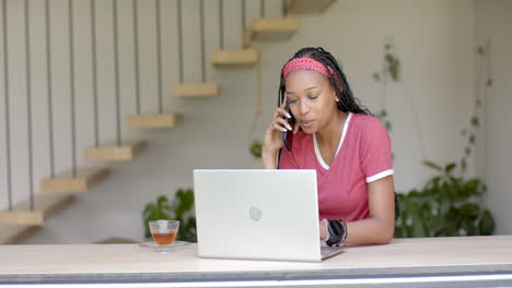 African-American-young-woman-talking-on-phone,-working-on-laptop-at-home