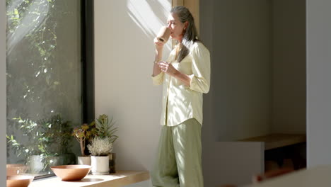 Caucasian-mature-woman-standing-by-window,-holding-cup