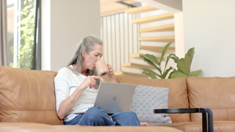 Mature-Caucasian-woman-sitting-on-sofa,-drinking-coffee-and-looking-at-laptop
