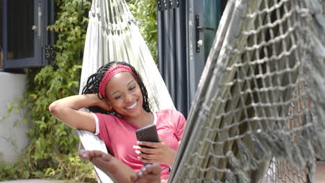 African-American-young-woman-relaxing-in-hammock,-holding-smartphone