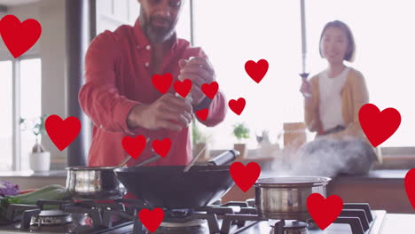 Animation-of-hearts-over-diverse-couple-embracing-cooking-in-kitchen