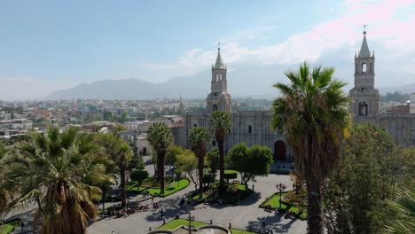 Stunning-flight-over-Arequipa's-Plaza-de-Armas,-among-palm-trees,-unveiling-towers-and-cathedral,-with-volcanoes-in-the-background