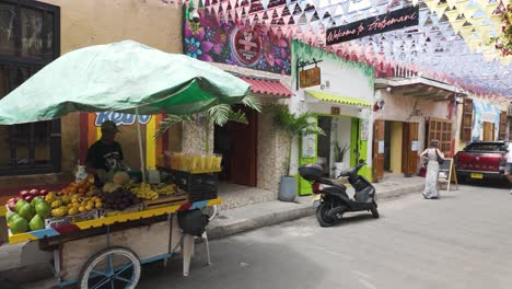 Colorful-fruit-cart-on-a-vibrant-street-in-Getsemani,-Cartagena,-with-decorative-flags-and-local-shops