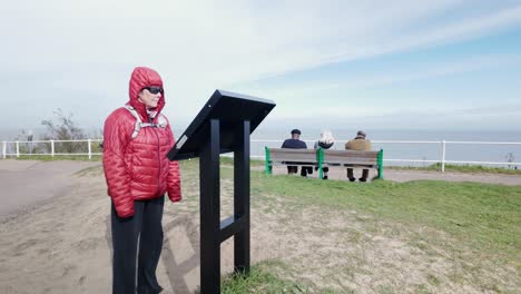 Female-coastal-walker-reads-info-board,-Suffolk-seaside-resort