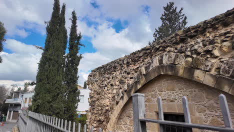 A-close-up-of-a-weathered-stone-arch-and-wall-in-Nicosia,-Cyprus,-with-tall-cypress-trees-in-the-background