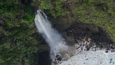 Tall-waterfall-spews-off-into-black-rock-cove-on-coastal-tropical-island-with-waves-crashing