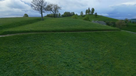 Lush-green-landscape-with-rolling-hills,-a-winding-path,-and-scattered-buildings-under-a-cloudy-sky