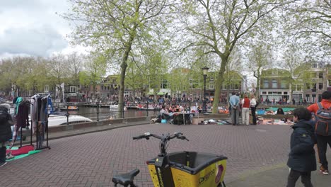 People-browsing-market-stalls-by-a-canal-during-King's-Day-in-Utrecht,-Netherlands