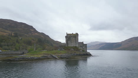 Aerial-flying-over-water,-circling-Eilean-Donan-Castle-on-cloudy-day,-Scotland