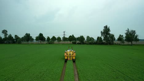 Blue-tractor-with-a-folded-yellow-field-sprayer-drives-through-a-green-grain-field